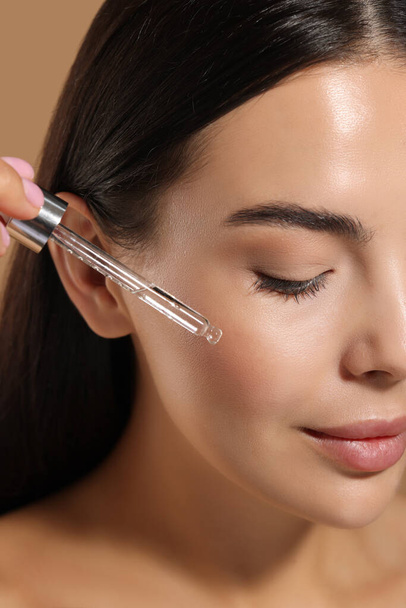 Beautiful young woman applying serum onto her face on beige background, closeup - Zdjęcie, obraz