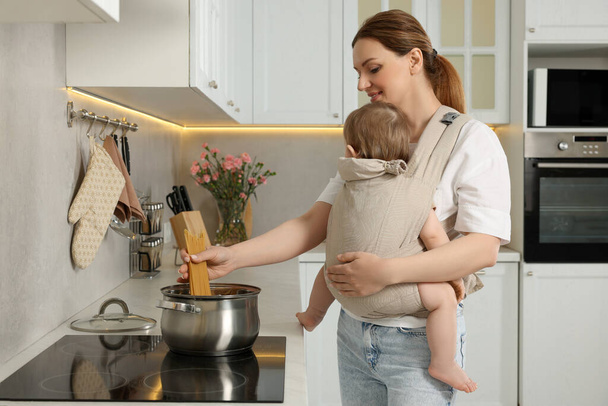 Mother holding her child in sling (baby carrier) while cooking pasta in kitchen - Фото, изображение