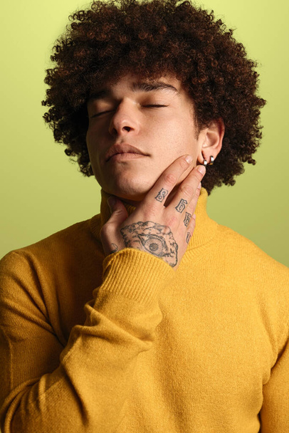 Calm young Hispanic male millennial with dark curly hair and tattoo on hand in yellow turtleneck touching neck with closed eyes against green background - Fotó, kép