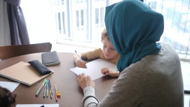 Assistere una madre musulmana in un hijab che guida i suoi figli di 6 e 8 anni attraverso i loro compiti scolastici a casa, mescolando famiglia educatio - Filmati, video