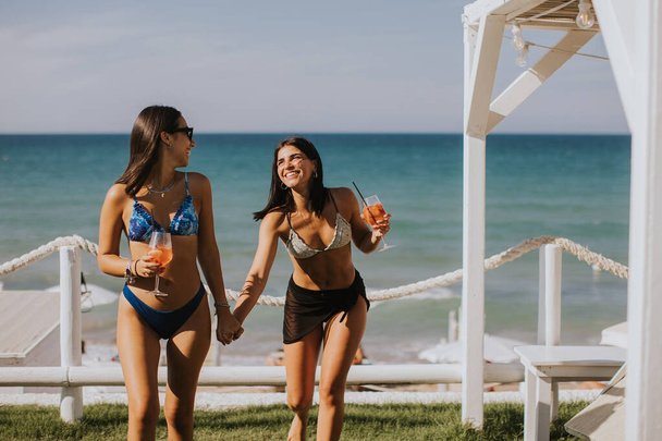 Két mosolygó fiatal nő bikiniben élvezi a nyaralást a tengerparton, miközben koktélt iszik. - Fotó, kép