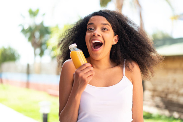 Junge Afroamerikanerin hält mit Überraschung und schockiertem Gesichtsausdruck Orangensaft im Freien - Foto, Bild