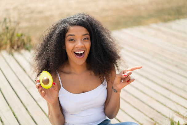 Jovem afro-americana segurando um abacate ao ar livre surpreso e apontando o dedo para o lado - Foto, Imagem