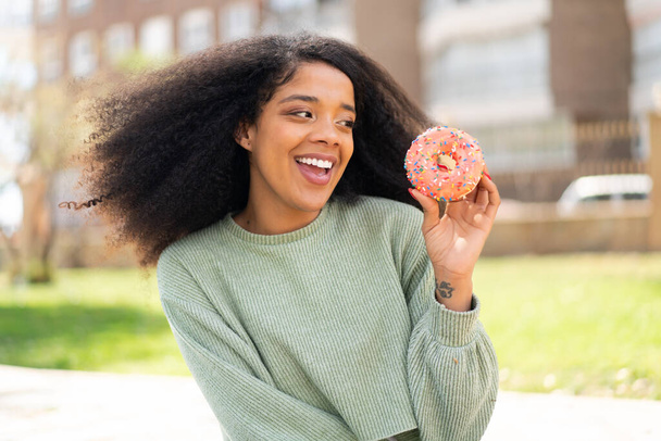 若いアフリカ系アメリカ人女性がドーナツをアウトドアに持ち込んで幸せな表情で - 写真・画像