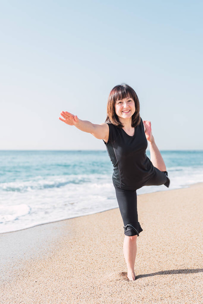  mujer en ropa deportiva equilibrio en la pierna en Natarajasana. en la estera mientras hace yoga en la costa arenosa cerca del mar en el día soleado y mirando a la cámara - Foto, imagen