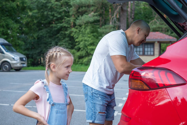 Egy copfos lány áll érdekesen az apja mellett egy piros autó csomagtartójában, egy parkolóval a háttérben. Kiváló minőségű fénykép - Fotó, kép