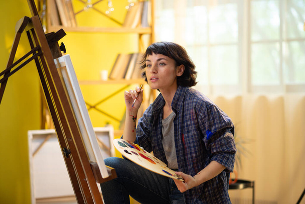 A beautiful woman with a funky pixie haircut is painting something on a large canvas and wearing a blue flannel top that has paint marks on it while being in an art studio. Portrait - 写真・画像