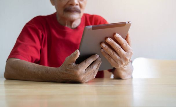Jóképű, piros pólós öregember digitális tabletet használ, miközben otthon dolgozik a kamerába néz. Szelektív fókusz. - Fotó, kép