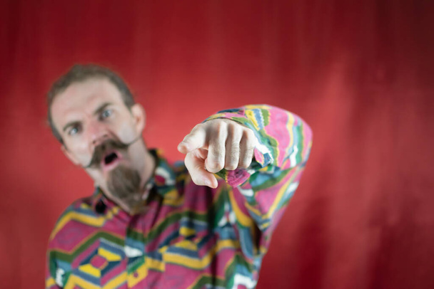 Homme hipster en colère pointant vers la caméra en criant et avec une expression furieuse avec fond rouge - Photo, image
