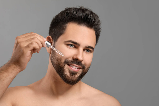 Handsome man applying cosmetic serum onto face on grey background, space for text - Фото, изображение