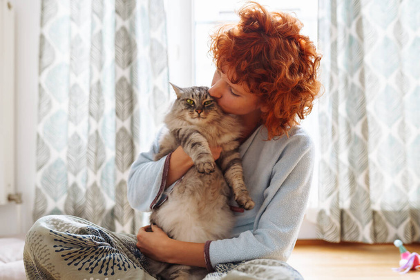 Funny red-haired girl teenager sitting on floor near window at home hugs kisses a purring Maine Coon cat. Friendship between pet and its owner - Photo, Image