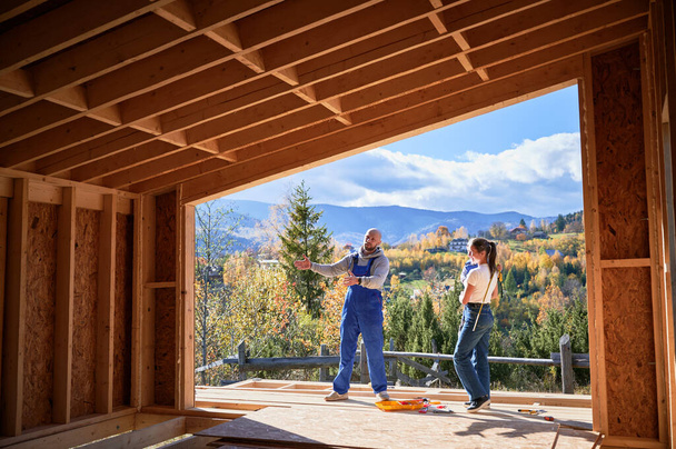 Otec, matka a syn staví dřevěný rámový dům. Muž v modrých montérkách ukazuje budoucí domov, stojí na terase na staveništi. Tesařství, rodinný koncept. - Fotografie, Obrázek