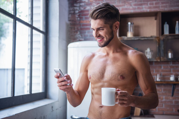 Σέξι νεαρό άνδρα με γυμνό τον κορμό εκμετάλλευση από ένα φλιτζάνι, χρησιμοποιώντας ένα smartphone και χαμογελώντας, ενώ στέκεται στην κουζίνα στο σπίτι - Φωτογραφία, εικόνα