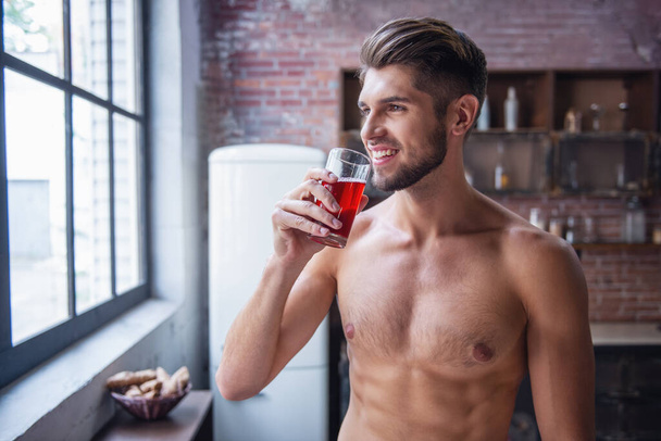 Sexy joven con torso desnudo está bebiendo jugo, mirando por la ventana y sonriendo mientras está de pie en la cocina en casa
 - Foto, Imagen