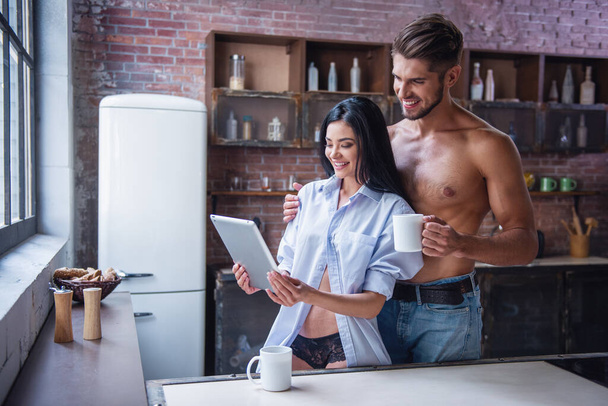 Belo jovem casal está bebendo café, usando um tablet digital e sorrindo enquanto está de pé na cozinha em casa
 - Foto, Imagem