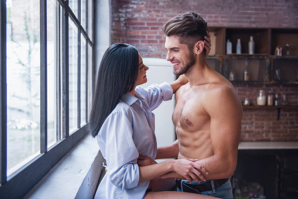 Beautiful young couple is hugging and smiling while standing in kitchen at home - Photo, Image