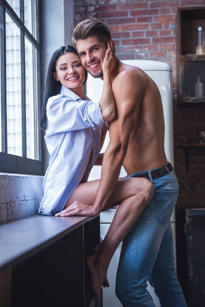 美しい若い情熱的なカップルがカメラを見て、自宅の台所でハグしながら笑みを浮かべて - 写真・画像