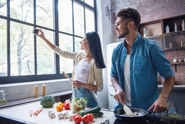 美しい若いカップルが selfie スマート フォンを使って、自宅のキッチンで料理をしながら笑顔をやっています。 - 写真・画像