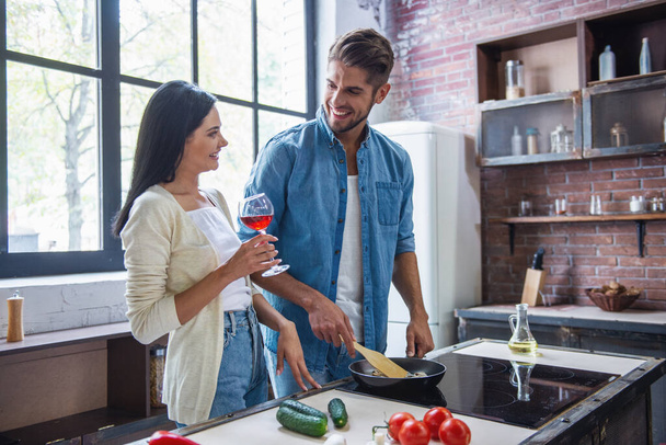 Krásná mladá dvojice se mluví a usmíval se při vaření v kuchyni doma. Žena pije víno, zatímco její muž je smažení potravin - Fotografie, Obrázek