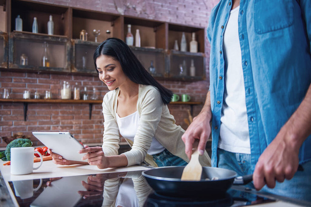 Όμορφη νεαρή γυναίκα είναι χρησιμοποιώντας ένα ψηφιακό tablet και χαμογελώντας, ενώ τον άντρα είναι το μαγείρεμα στην κουζίνα στο σπίτι - Φωτογραφία, εικόνα