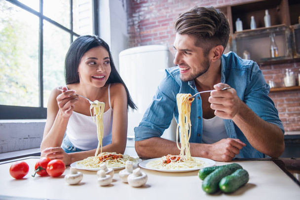Belo jovem casal está falando, olhando um para o outro e sorrindo enquanto come na cozinha em casa
 - Foto, Imagem
