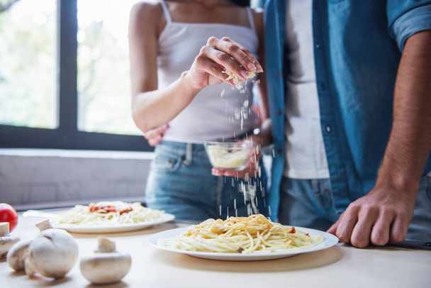 abgeschnittenes Bild eines schönen jungen Paares, das zu Hause in der Küche Spaghetti isst. Frau fügt geriebenen Käse hinzu - Foto, Bild