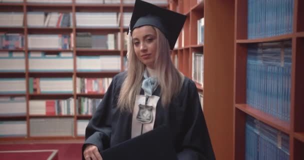 Απόφοιτη φοιτήτρια Περήφανα ανυψώνει το δίπλωμά της στην αίθουσα της πανεπιστημιακής βιβλιοθήκης. Έννοια πανεπιστημιακής εκπαίδευσης - Πλάνα, βίντεο