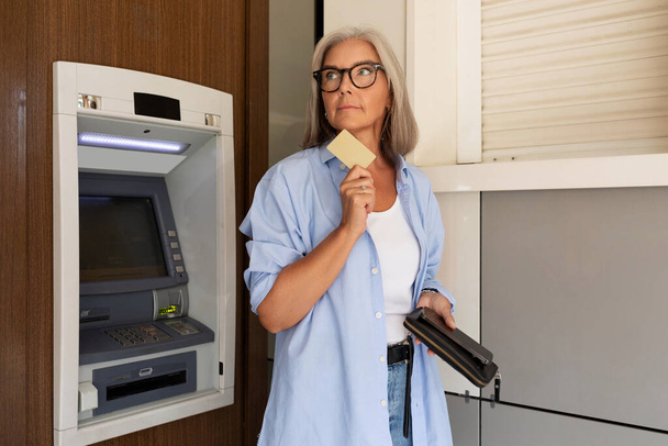 žena středního věku stojí v létě u bankomatu a drží peněženku. - Fotografie, Obrázek