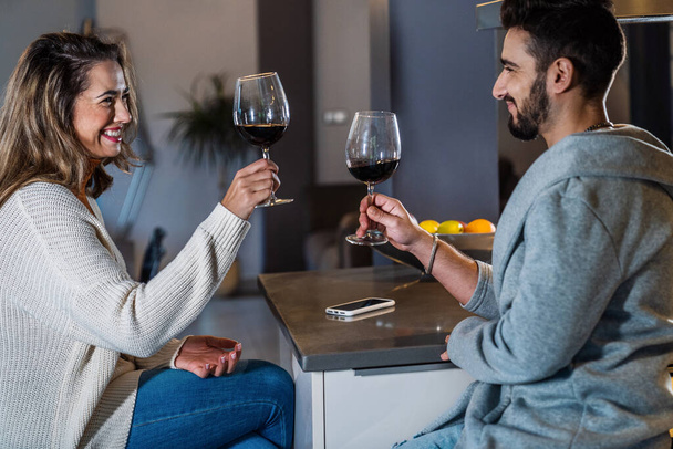 Τρόπος ζωής δύο νέων ανθρώπων με ένα ποτήρι κρασί σε ένα ρομαντικό και χαρούμενο βράδυ στην κουζίνα του αγροτικού τους σπιτιού. Κοντινό πλάνο ενός ζευγαριού. Ευτυχισμένο ζευγάρι που κάνει πρόποση με ένα ποτήρι κόκκινο κρασί. - Φωτογραφία, εικόνα