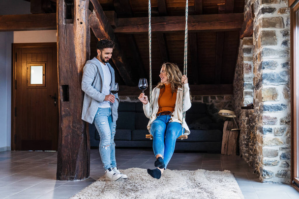 Un couple prenant un verre de vin sur une balançoire installée dans le grenier de leur maison rurale, style de vie d'un jeune couple lors d'une soirée romantique et heureuse. - Photo, image