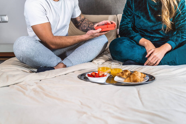 Lähikuva nuoren parin aviomiehestä, jota ei voida tunnistaa pyjamassa, antaa lahjan vaimolleen, koska he syövät aamiaista sängyssä nykyaikaisen kodin makuuhuoneessa. Vuosijuhlaa. Romanttinen aamiainen. Kotimainen elämäntapa. - Valokuva, kuva