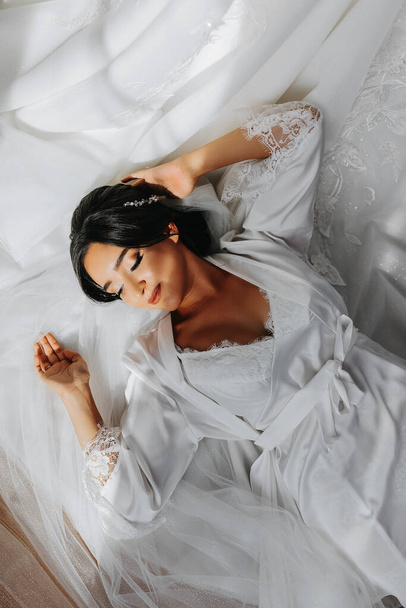 Portrait of a girl in a morning robe. Happy bride with professional make-up and long veil posing for photographer. The bride is preparing for the wedding - Фото, изображение