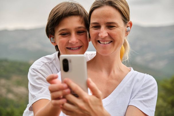 Retrato de la madre y el hijo feliz en ropa casual sonriendo mientras toma selfie en el teléfono inteligente y disfrutar de vacaciones en un fondo borroso de la naturaleza - Foto, Imagen