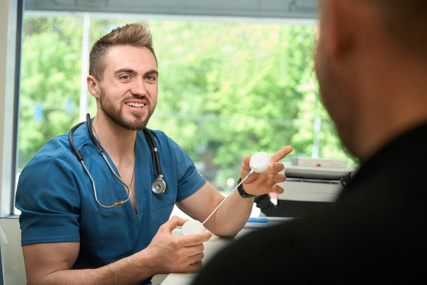 Gülümseyen genç fizyoterapist, elektromiyomülasyon seansından önce hastasına elektrot pedlerini gösteriyor. - Fotoğraf, Görsel