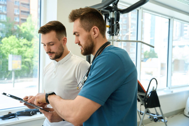 Physiotherapeut zeigt auf Papier am Klemmbrett zum Reha-Trainer im Fitnessstudio - Foto, Bild