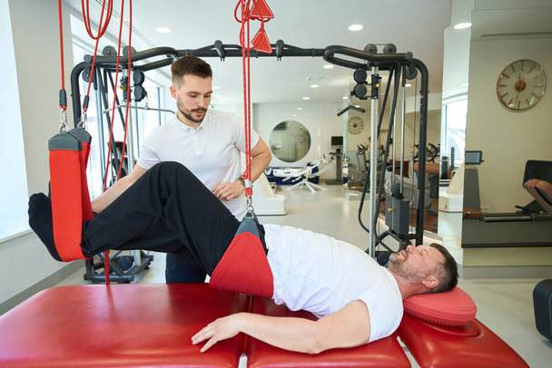 Homme adulte faisant flexion du genou couchée sur un entraîneur de suspension assisté par un physiothérapeute - Photo, image