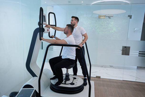 Man seated on stool of multi-axis motorized platform doing balance exercise supervised by experienced physiotherapist on machine touchscreen - Photo, image