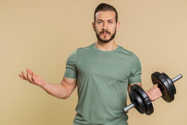 Athlete sportive man showing indignant expression, asking why, what reason of failure, demonstrating disbelief irritation by troubles. Middle eastern sportsman guy with dumbbell on beige background - Photo, Image