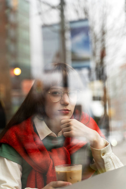 Una bella donna pensierosa con gli occhiali seduta al bar, che guarda la finestra, che tiene in mano da bere. Donna seria, pausa caffè mattutina - Foto, immagini