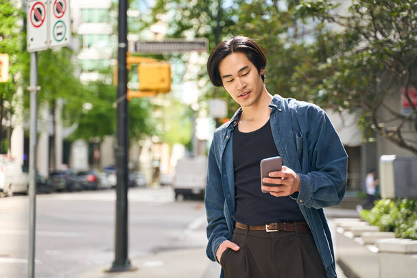 Giovane uomo asiatico sorridente che tiene smartphone tramite app mobile, lettura di messaggi di testo, comunicazione online camminando per strada urbana. Giapponese hipster ragazzo shopping online all'aperto - Foto, immagini