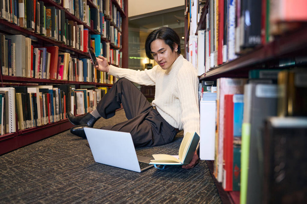 Χαλαρός Ιάπωνας σπουδάζει φορητό υπολογιστή, κρατάει χάρτινο βιβλίο και smartphone, προετοιμάζεται για εξετάσεις καθισμένος στη βιβλιοθήκη του πανεπιστημίου, εκπαίδευση. Ασιατικές γλώσσες μάθησης σπουδαστών - Φωτογραφία, εικόνα