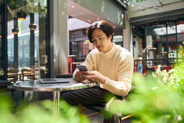 Hezký usměvavý Asiat držící mobilní telefon, kontrolující e-mail, čtoucí textovou zprávu. Mladý sebevědomý japonský podnikatel pomocí notebooku pracující on-line sedí v moderní kavárně. Technologický koncept - Fotografie, Obrázek