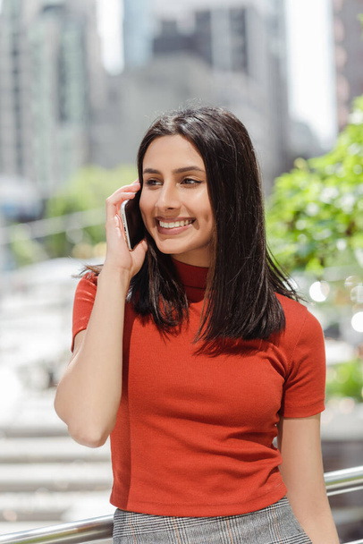 Authentic portrait of smiling beautiful Indian woman talking on mobile phone, answering call looking away standing on urban street, copy space. Technology concept  - Photo, image