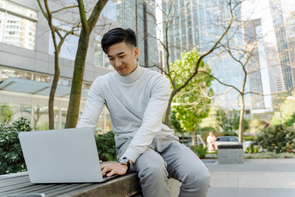 Привлекательный азиатский мужчина с ноутбуком, сидит на скамейке на открытом воздухе. Китайский бизнесмен работает в сети, удаленная работа. Привлекательный студент готовится к экзамену. Концепция онлайн образования - Фото, изображение