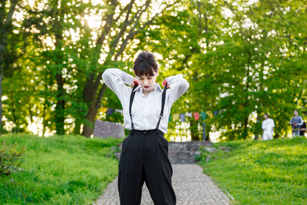 Красивая девушка в белой рубашке, в черных брюках с подтяжками на фоне неба и зеленой травы крупным планом - Фото, изображение