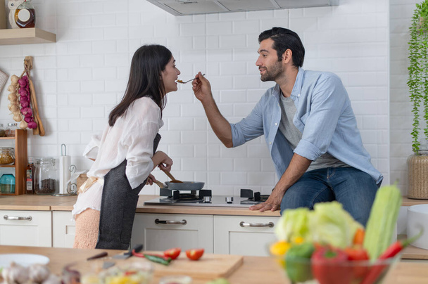 Ευτυχισμένο νεαρό ζευγάρι που μαγειρεύουν μαζί στον πάγκο της κουζίνας, ταΐζοντας ο ένας τον άλλον στην κουζίνα τους στο σπίτι - Φωτογραφία, εικόνα