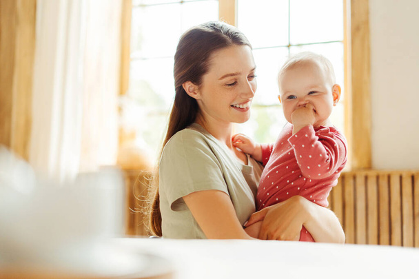 Young smiling mother holding her cute little baby sitting at comfortable home. Love, care, motherhood concept - Valokuva, kuva