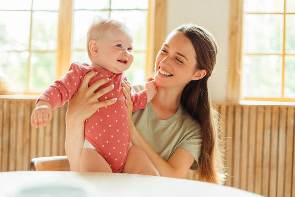 Young mom smiling at her little baby, holding hands, having leisure, time together. Positive female babysitter playing with little beautiful baby. Concept of children's leisure and wellbeing, parenthood - Photo, Image