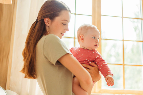 Gyönyörű mosolygós anya kezében kis aranyos baba piros ruhát a nappaliban otthon. Vonzó fiatal dadus gyerekkel. Szerelem, gondozás, szülőség fogalma - Fotó, kép