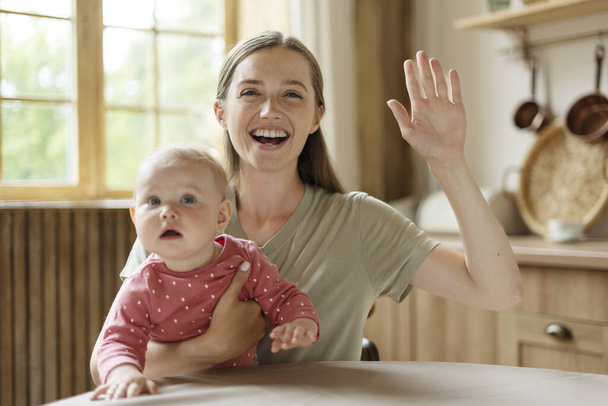 Lächelnde junge Mutter mit kleinem süßen Baby, das Videoanruf hat, winkende Hand, in der Küche sitzend, von zu Hause aus arbeitend, Fernarbeit. Schöne weibliche Video anschauen. Konzept der Kommunikation, Mutterschaft  - Foto, Bild